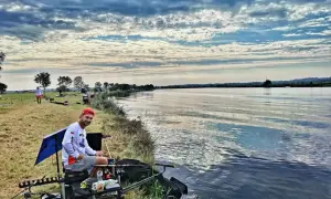 Mężczyzna w czerwonej czapce siedzi nad wodą, gotowy do łowienia ryb metodą picker feeder. W tle widać innych wędkarzy i piękne, chmurne niebo.