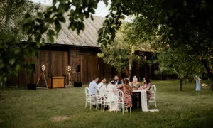 Goście weselni siedzą przy stole na trawie, w tle drewniana stodoła. To urokliwa brzozowa wyspa podkrajewo, gdzie odbywa się przyjęcie.