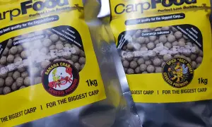 Dwie paczki z żółtymi etykietami zawierają kulki na karpia. Na opakowaniach widnieje napis "CarpFood" i grafika z pszczołą.