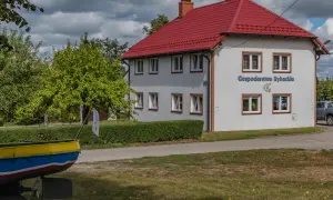 Budynek z czerwonym dachem, na ścianie napis "Gospodarstwo Rybackie" i ryba. Obok łódź i zielone drzewa.