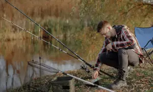 Mężczyzna przygotowuje się do łowienia na grunt nad jeziorem, zakładając przynętę na haczyk. W tle wędki i podbierak.