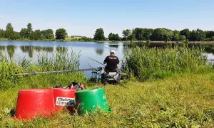 Wędkarz z napisem "FEBI FISH" na koszulce siedzi nad spokojnym stawem, gotowy do łowienia.