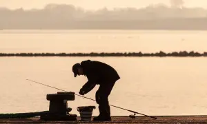 Wędkarz przygotowuje zanętę do koszyczka, stojąc nad wodą o wschodzie słońca.