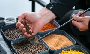 Wędkarz przygotowuje zanętę, trzymając w dłoni pelet. W tle widać pojemniki z różnymi składnikami, w tym kukurydzą.