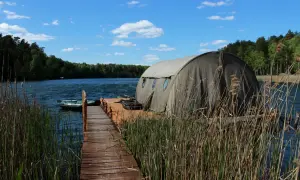 Drewniany pomost prowadzi do namiotu na wodzie, idealnego na łowisko karpiowe Smolno. W tle las i błękitne niebo.