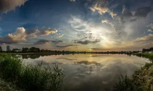 Zachód słońca nad jeziorem, odbijający się w wodzie. W oddali widać drzewa i zabudowania, a na pierwszym planie rośnie trawa. Widok pod borem ochaby.