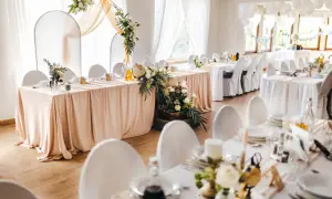 Elegancka sala weselna z białymi obrusami i kwiatami. Na stole pod dębem przywory stoi globus i walizka.