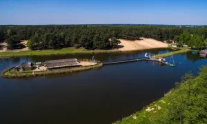 Malowniczy widok na jezioro z pomostami i budynkami zadaszonymi strzechą. W tle las i piaszczysta plaża. Młynek w Tuszynie to idealne miejsce na wypoczynek.