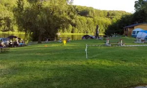 Relaksacyjny camping nad stawami. Ludzie siedzą przy stołach, w tle drzewa i woda. W oddali widać domek i namioty.