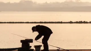 Wędkarz przygotowuje zanętę do koszyczka, stojąc nad wodą o wschodzie słońca.