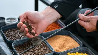 Wędkarz przygotowuje zanętę, trzymając w dłoni pelet. W tle widać pojemniki z różnymi składnikami, w tym kukurydzą.