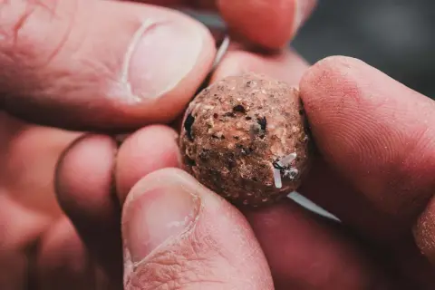 Dłonie trzymają kulkę pelletu z haczykiem. Prosty sposób, jak założyć pellet na haczyk, by łowić ryby.