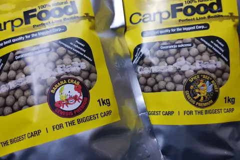 Dwie paczki z żółtymi etykietami zawierają kulki na karpia. Na opakowaniach widnieje napis "CarpFood" i grafika z pszczołą.