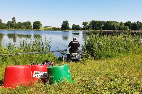 Wędkarz z napisem "FEBI FISH" na koszulce siedzi nad spokojnym stawem, gotowy do łowienia.