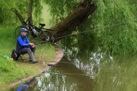 Wędkarz siedzi nad wodą, czekając na branie. Obok rower, a w zanęcie pellet na suma.
