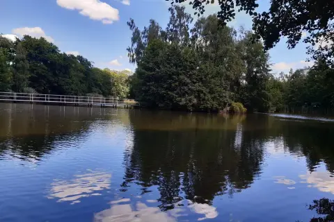 Spokojne jezioro z odbijającymi się chmurami i drzewami. W oddali biały mostek, idealne miejsce na wypoczynek w Golina ośrodek wypoczynkowy.