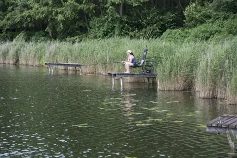 Kobieta siedzi na pomoście, łowiąc ryby. Wokół trzciny i las. Idealne łowisko rybie oko.