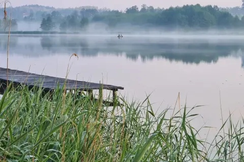 Poranna mgła nad zalewem w Janowicach. Drewniany pomost i trzciny na pierwszym planie, na wodzie mała łódka.