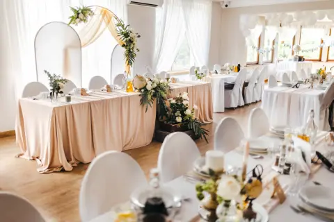 Elegancka sala weselna z białymi obrusami i kwiatami. Na stole pod dębem przywory stoi globus i walizka.