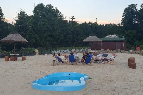 Relaks na piasku w Krężnicy Jarej. Grupa przyjaciół odpoczywa na leżakach przy małym basenie, w tle drewniana sauna i palmy.