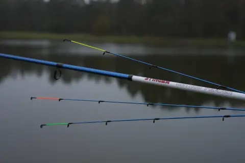Wędki Robinson Stinger Method Feeder z kolorowymi szczytówkami na tle spokojnej wody i drzew.