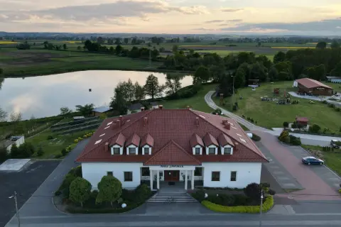 Nasz dworek w Dzierżowicach, otoczony zielenią i stawem, zaprasza do odpoczynku. W tle rozciągają się pola i pagórki.