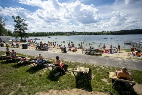 Tłum ludzi odpoczywa na plaży nad jeziorem Kierskim Małym. Słońce świeci, a chmury tworzą malowniczy krajobraz.