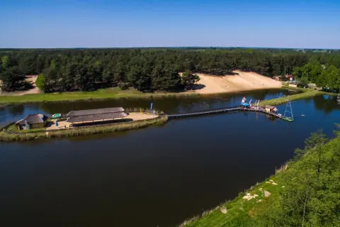 Malowniczy widok na jezioro z pomostami i budynkami zadaszonymi strzechą. W tle las i piaszczysta plaża. Młynek w Tuszynie to idealne miejsce na wypoczynek.