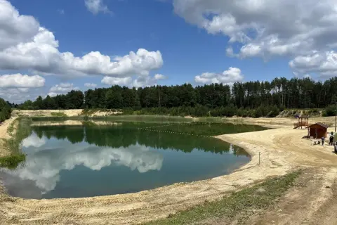 Zbiornik Czarna Sędziszowska z piaszczystą plażą i lasem w tle. Błękitne niebo z białymi chmurami odbija się w spokojnej tafli wody.