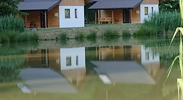 Urokliwe łowisko z domkami w Podkarpackiem. Dwa drewniano-białe domki z dachami odbijają się w spokojnej tafli wody.