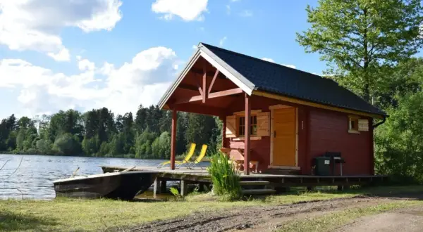 Urokliwe łowisko z domkami na Mazowszu. Czerwony domek z werandą, łódką przy brzegu i dwoma żółtymi leżakami zaprasza do relaksu nad jeziorem.