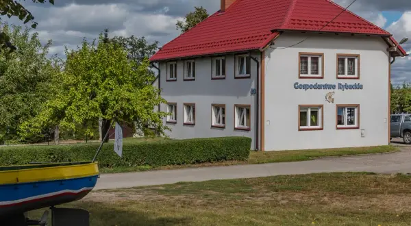 Budynek z czerwonym dachem, na ścianie napis "Gospodarstwo Rybackie" i ryba. Obok łódź i zielone drzewa.