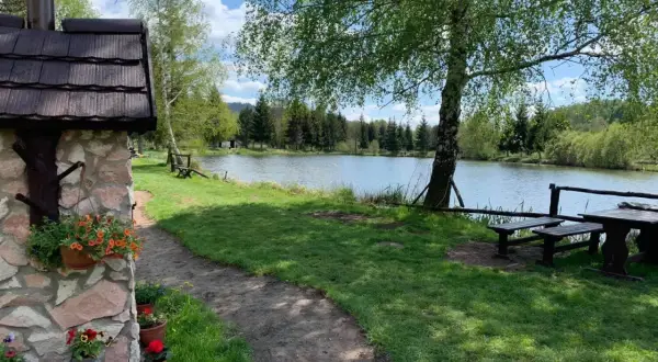 Kamienny domek z kwiatami w doniczkach, ścieżka prowadząca nad potokiem grzędy, ławki i stół pod drzewem.