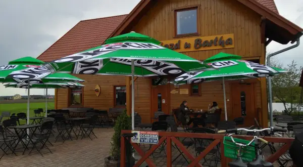 Drewniany budynek z zielonymi parasolami Browaru Okocim. Stoliki i krzesła na zewnątrz. U Bartka w Miliczu.