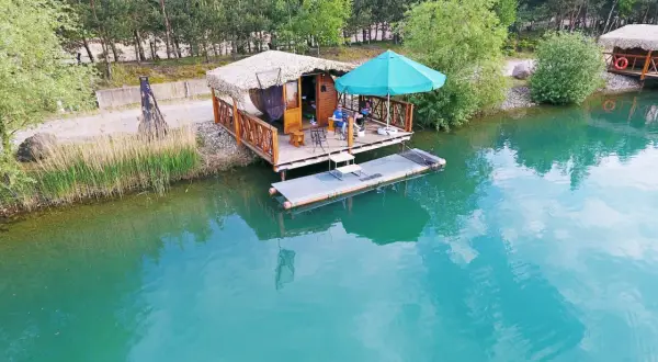 Relaks nad turkusowym zalewem Dobroszyce. Drewniana chata na wodzie, parasol i wędkarze.
