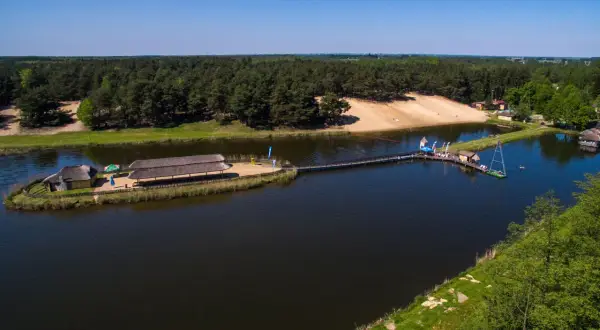 Malowniczy widok na jezioro z pomostami i budynkami zadaszonymi strzechą. W tle las i piaszczysta plaża. Młynek w Tuszynie to idealne miejsce na wypoczynek.