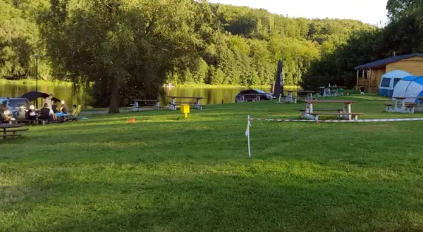 Relaksacyjny camping nad stawami. Ludzie siedzą przy stołach, w tle drzewa i woda. W oddali widać domek i namioty.