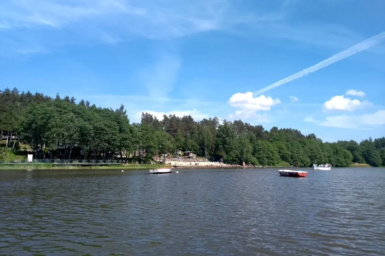 Letnie popołudnie nad jeziorem Osuszyno. Woda spokojna, na brzegu plażowicze, a na jeziorze łodzie. Może nawet jakieś ryby pływają pod powierzchnią.