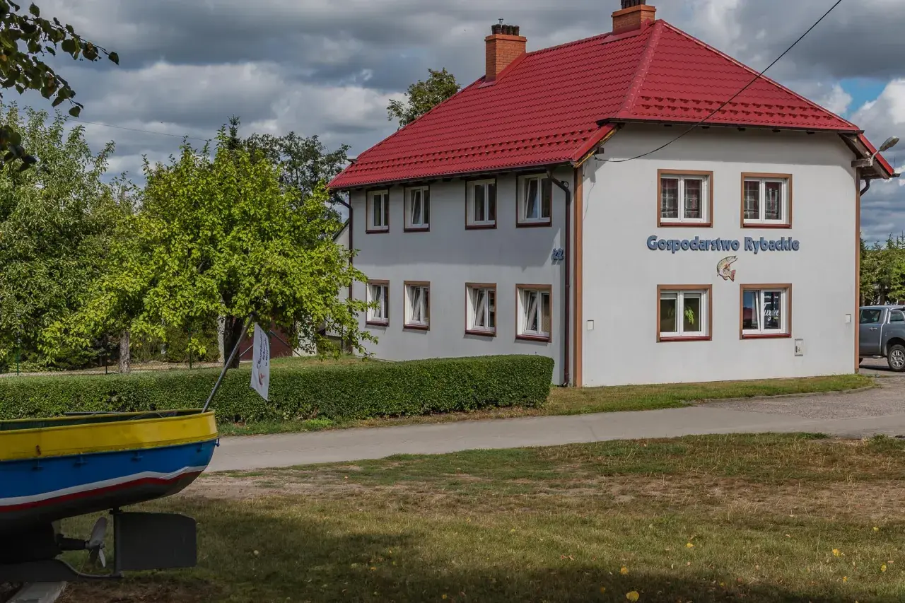 Budynek z czerwonym dachem, na ścianie napis "Gospodarstwo Rybackie" i ryba. Obok łódź i zielone drzewa.