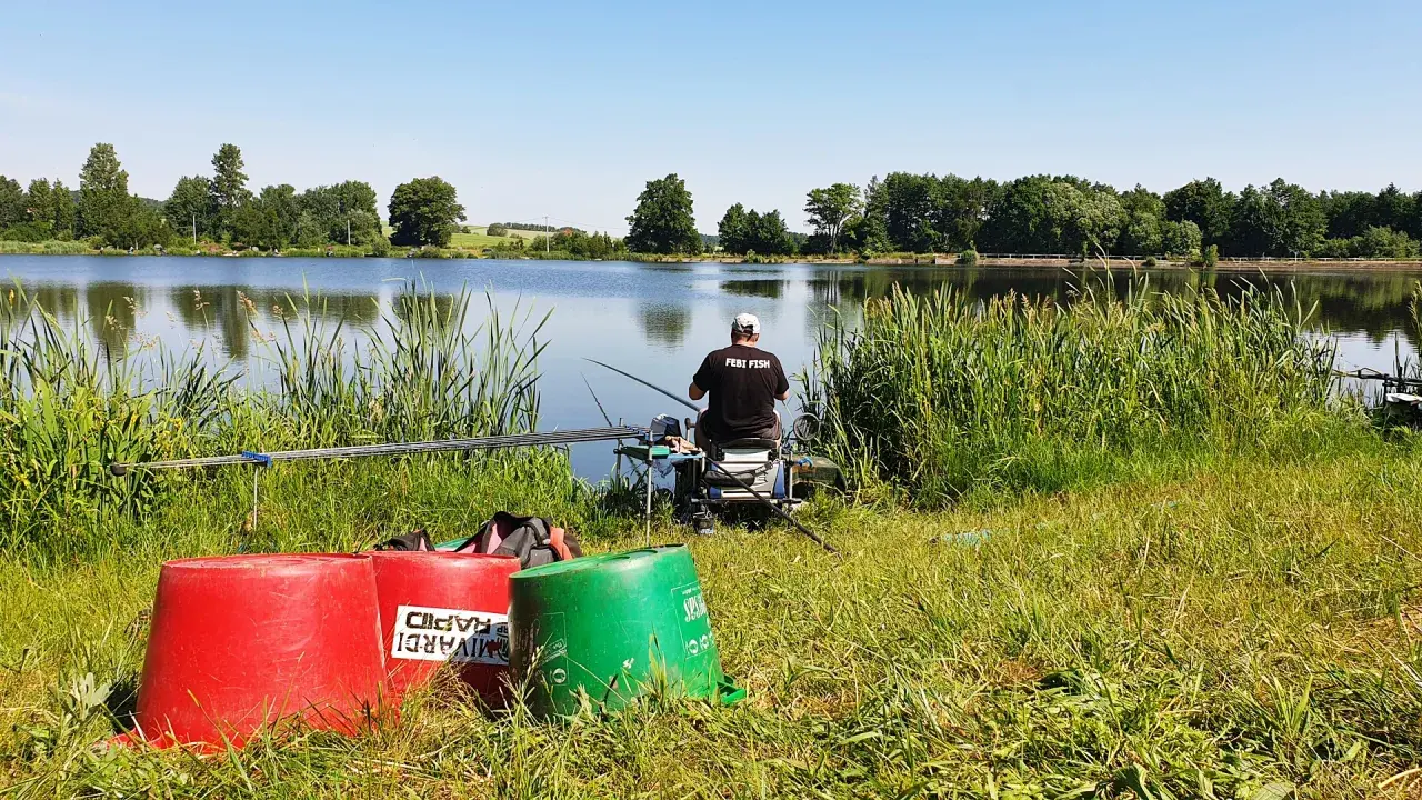 Wędkarz z napisem "FEBI FISH" na koszulce siedzi nad spokojnym stawem, gotowy do łowienia.