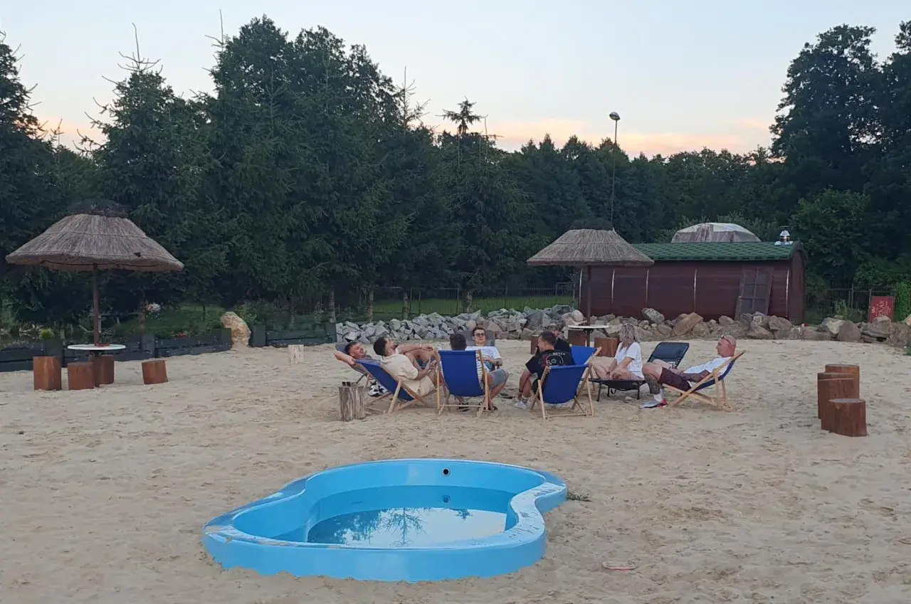Relaks na piasku w Krężnicy Jarej. Grupa przyjaciół odpoczywa na leżakach przy małym basenie, w tle drewniana sauna i palmy.