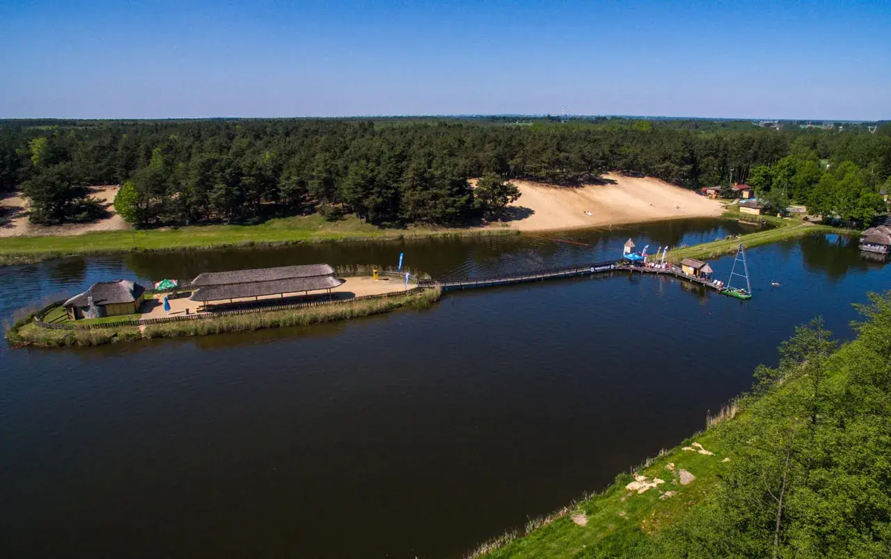 Malowniczy widok na jezioro z pomostami i budynkami zadaszonymi strzechą. W tle las i piaszczysta plaża. Młynek w Tuszynie to idealne miejsce na wypoczynek.