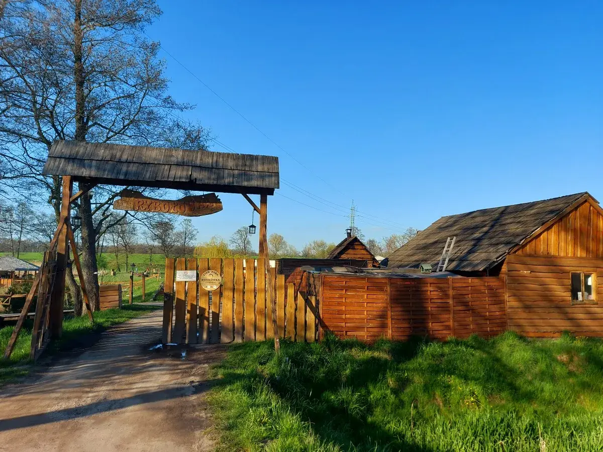 Drewniana brama z napisem "RYBOŁÓWKA" prowadzi do obiektu w Mikołowie. W tle widać chaty i zielone łąki.