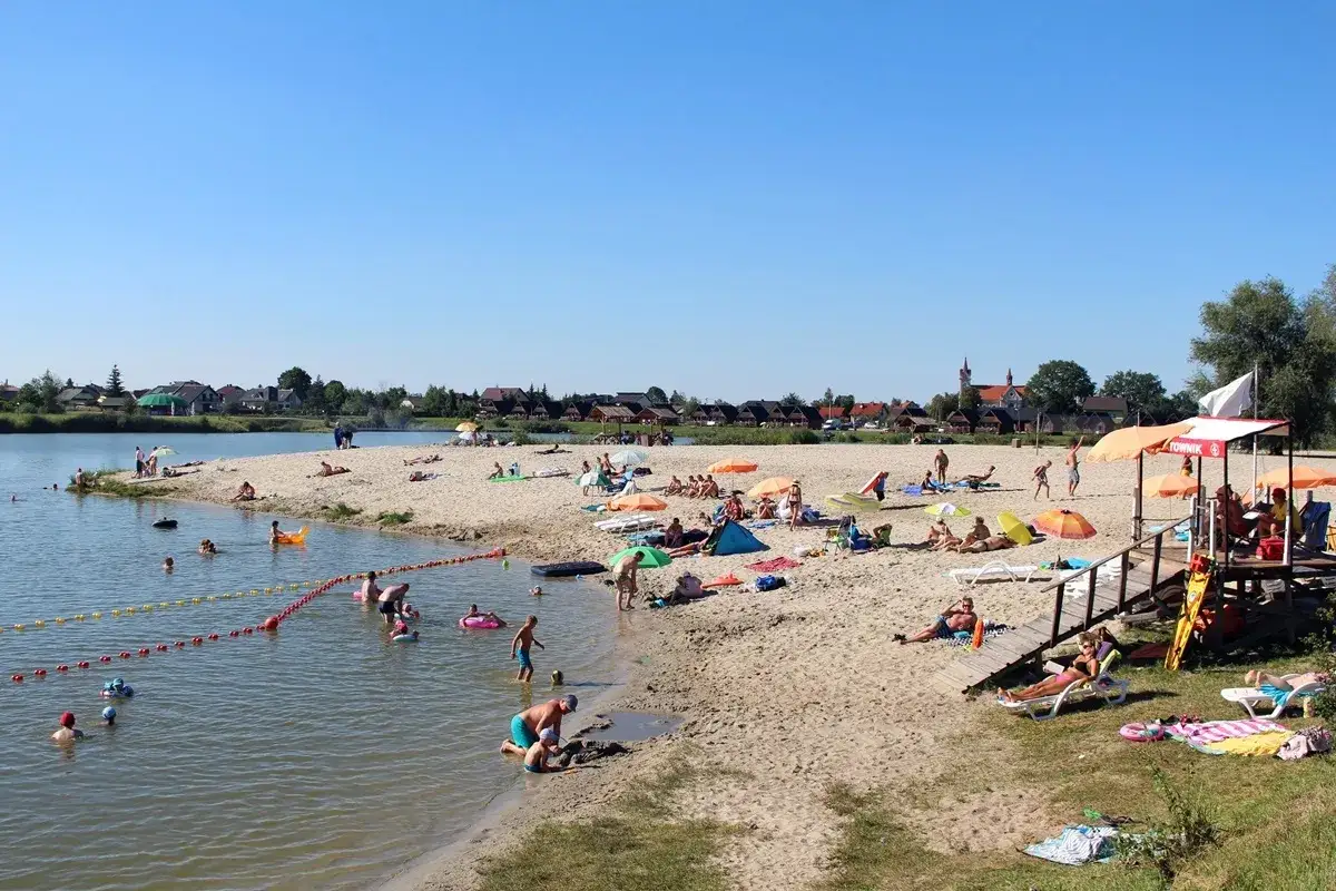Słoneczny dzień na piaszczystej plaży bobrowego rozlewiska w Zabierzowie Bocheńskim. Ludzie odpoczywają, kąpią się i bawią w wodzie.
