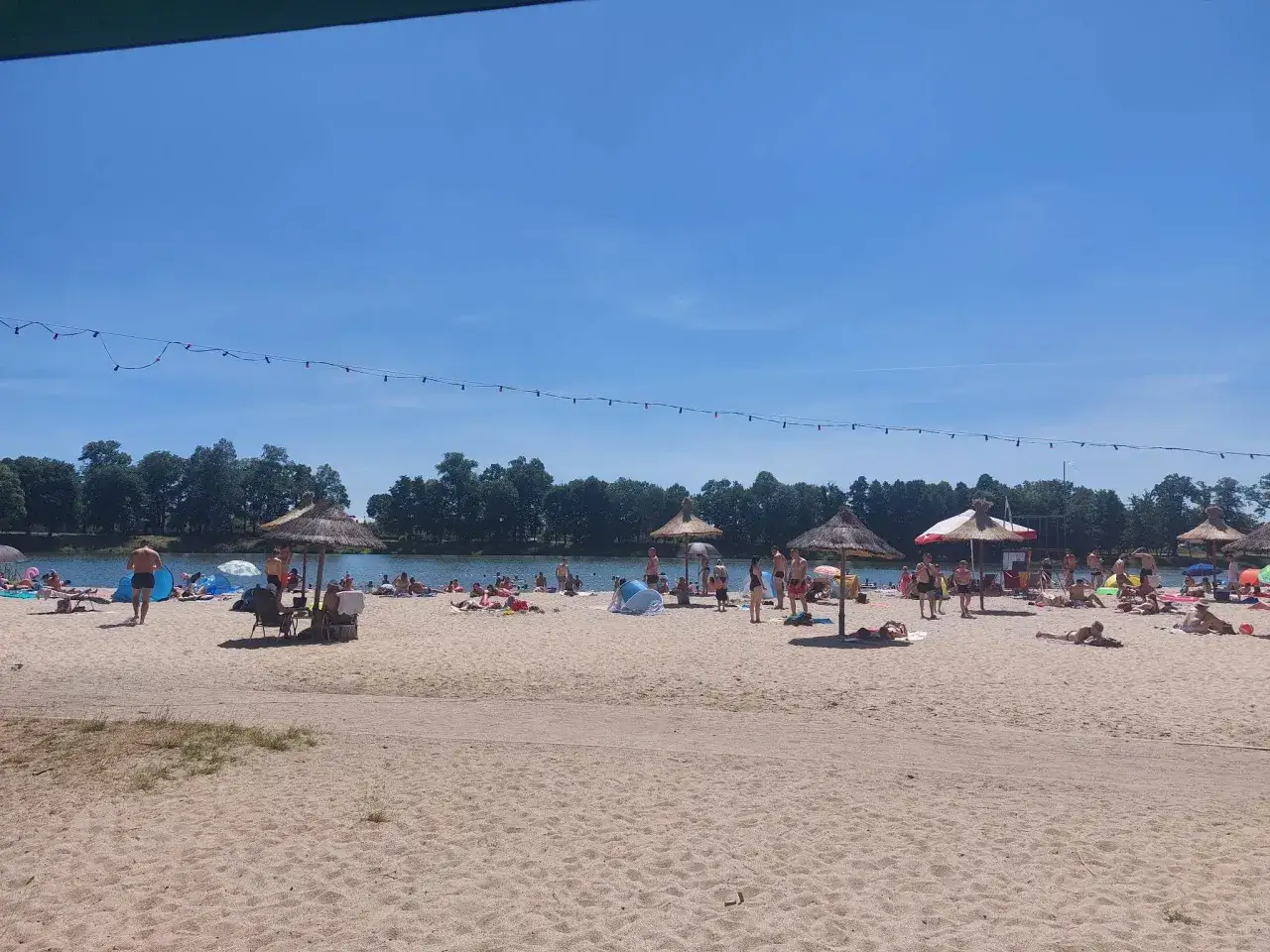 Słoneczny dzień na plaży nad zalewem Małomice. Ludzie odpoczywają na piasku, kąpią się i opalają.