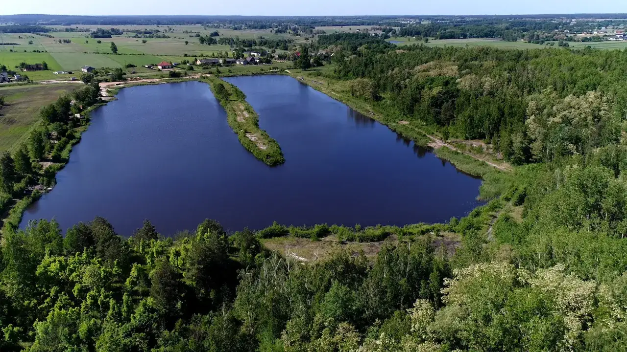Malownicze łowisko borowy młyn otoczone zielenią. Spokojna tafla wody odbija błękit nieba, a wyspa pośrodku dodaje uroku.