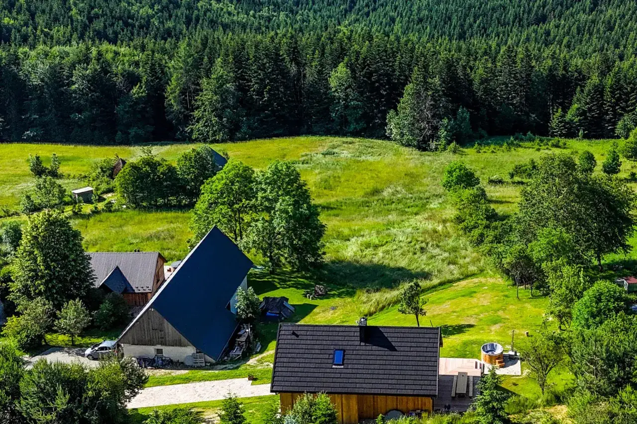 Chata na skraju lasu, otoczona zielenią. Widać kilka budynków, w tym dom z bali i drewnianą balię.