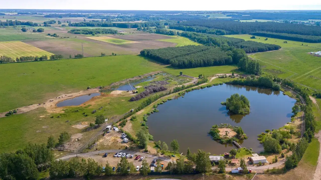 Malownicze łowisko wędkarskie Siedlnica z wyspą pośrodku, otoczone polami i lasami.