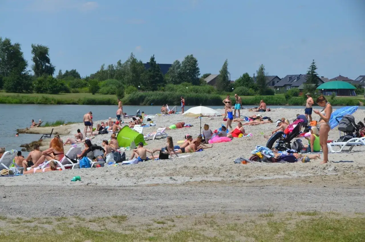 Ludzie odpoczywają na piaszczystej plaży nad wodą. W tle drzewa i zabudowania. To popularne bobrowe rozlewisko w Zabierzowie Bocheńskim.