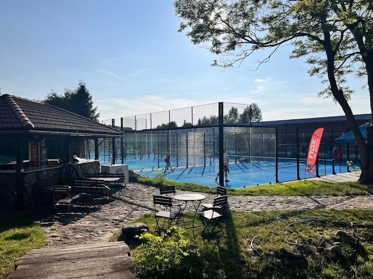 Słoneczny dzień na kortach padelowych w Rancho Mała Nieszawka. W tle widać niebieskie boiska otoczone siatkami, a z przodu zadaszona wiata z ławkami.